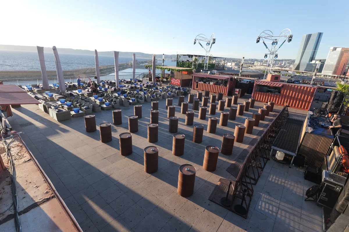 Wide view of the rooftop deck looking north-west over the Digue du Large breakwater and the port, drum tables in rows, container bars at the centre and the La Marseillaise tower on the right