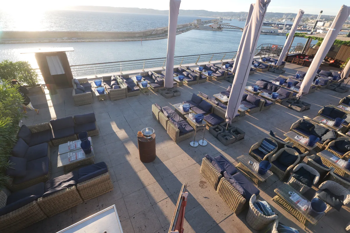 The sea-facing side of the rooftop at sunset, the sun low over the breakwater, rattan lounge seating with navy cushions and tall white closed parasols along the rail