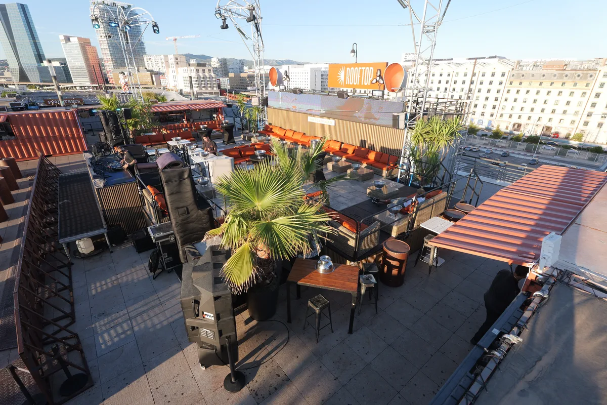The bar and stage corner of the rooftop, with a fan palm, orange banquettes, speaker stacks and the venue's orange ROOFTOP sign on the parapet, the Marseille skyline behind